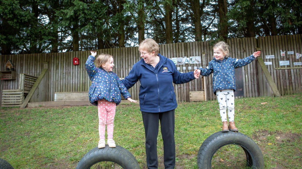 Older practitioner holding the hands of two young children standing on tyres in a nursery garden area