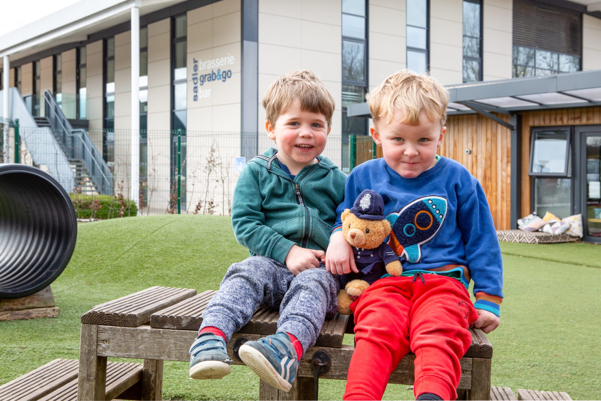 Two boys sat with each other outside of the nursery holding safety mascot PC Paws 